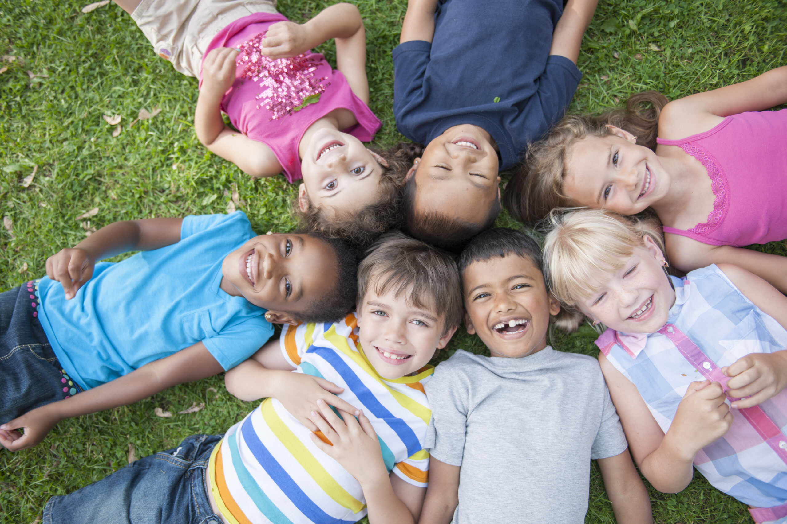 Lying in a Circle in the Grass A multi-ethnic group of elementary age children are lying in the grass at the park. They have their heads together in the middle of a circle and are smiling and looking up at the camera.